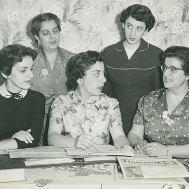 A black and white photo shows an image of five women together, two standing three sitting, engaged in conversation. In front of them is a spread of newspapers.
