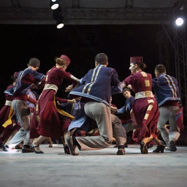 Young men and women, alternating, do a circle dance on stage in traditional costume.
