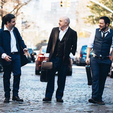 Three smiling men dressed in suits stand in the middle of a cobblestone street. They are in the midst of a conversation walking and holding their instruments in cases.