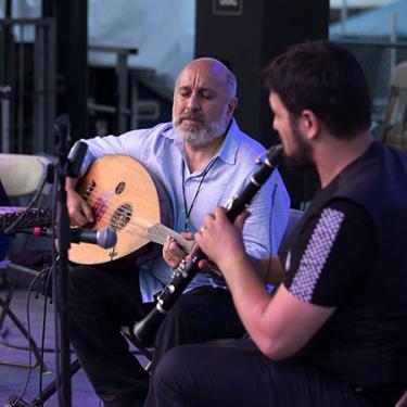 Under a purple light, a man in a white shirt closes his eyes as he strums the oud, an acoustic instrument with 11 strings. Next to him, a man with his back to the camera plays the clarinet.