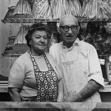 A black and white photo. Pictured are an elderly couple standing together in their bakery behind the counter.