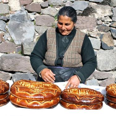 In front of a rock wall, a woman wearing a brown vest sits behind a table. On the table are placed several beautifully decorated circular loaves of bread.