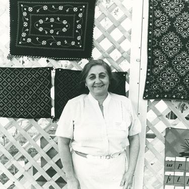 Depicted in black and white, an older woman stands smiling in front of a display of her impressively embroidered needlework.