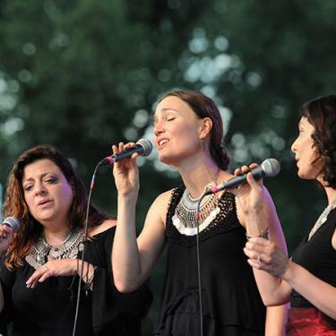 In front of dark green foliage, three women wearing black shirts and silver necklaces strand holding microphones. Eyes closed, they appear amid a performance.