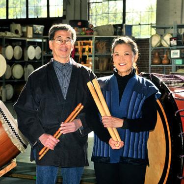 Two people each hold a pair of wooden beater sticks, standing among large taiko drums.