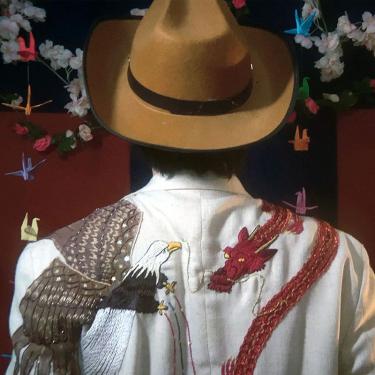 The back of a person wearing a brown fedora and white jacket embroidered with a bald eagle on the left shoulder fighting a red dragon on the right. Origami cranes are hanging in the background.