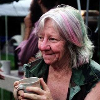 Carol Judy, with shoulder-length gray hair streaked with pink and blue, smiles and holds a mug.