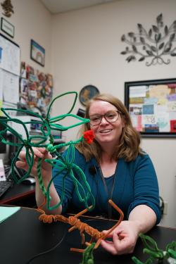 A woman wearing glasses holds up a ginseng plant made of pipe cleaners. The plant includes green leaves, red berries, and a brown root.  