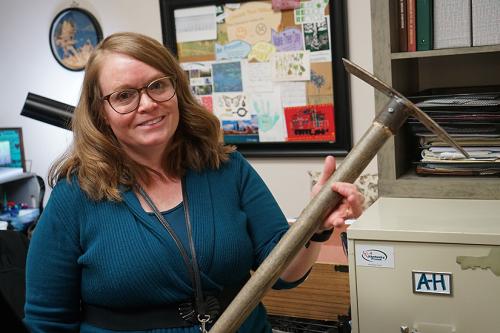 A woman wearing glasses smiles at the camera and holds up a pickaxe to her left, our right.  