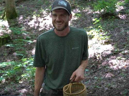 A man standing in the woods and wearing a baseball cap smiles at the camera. He is holding a basket in his left hand. 