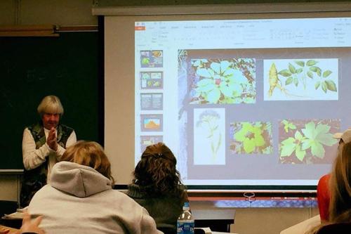 An elder woman speaks at the front of a classroom, next to a slide projection showing various plant photos.