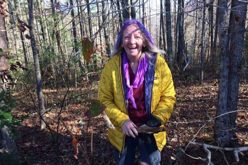 An elder woman in bright yellow jacket, standing in a woody area, laughs.