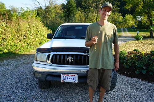 A smiling man wearing a baseball cap stands in front of a white Toyota car with an Ohio license plate that reads GINSENG. 