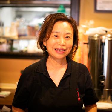 A woman in a black apron in the center of the frame smiles at the camera. The background, inside a restaurant, is slightly blurred.