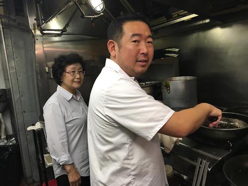 A man in an industrial kitchen holds a frying pan over a stove while looking at the camera over his right shoulder. A woman wearing glasses stands behind him to the left and smiles at the camera.  
