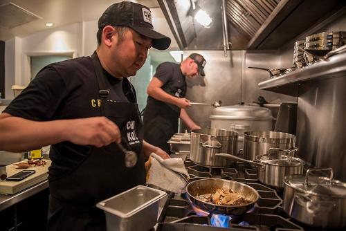 Two men wearing baseball hats standing cooking at an industrial stove. The man in the foreground is using a frying pan and the man is slightly blurry. 