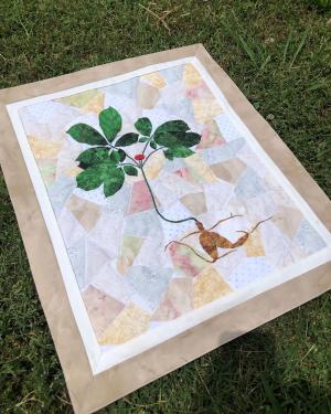 A quilted ginseng plant is laid out on grass. The background of the quilt is made up of patterned pastel fabric cut into geometric shapes. 