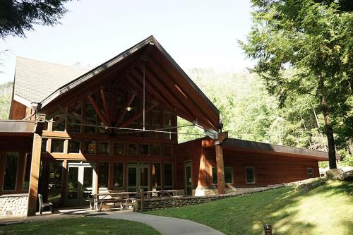 A single-story building with a large, two-story, central structure featuring wall to ceiling windows and a sloped roof. The rest of the building is wood paneled and there is outdoor seating on a small patio.