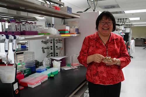 A woman wearing glasses smiles at the camera and holds a grown plant in her hand. To her right, our left, is a laboratory counter. 