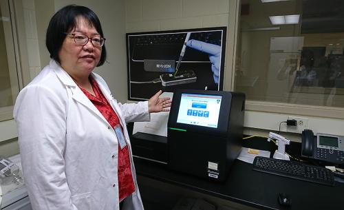 A woman wearing glasses and a white lab coat stands to the left of the frame and gestures to a computer screen on her left, our right.