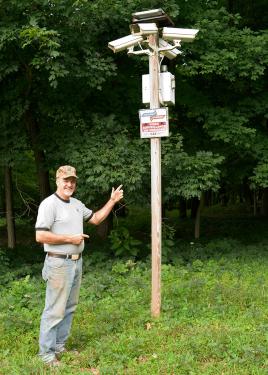 A man wearing a baseball cap smiles at the camera while pointing to the pole on his right, our left. The pole is crowned with four security cameras. 