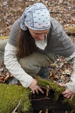 A woman wearing a bandana in her hair squats on the forest ground and lifts a moss-covered log in the foreground. 