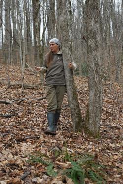 A woman wearing a bandana in her hair and black rubber boots stands in the forest facing the camera with her left arm hooked around a thin tree trunk. 