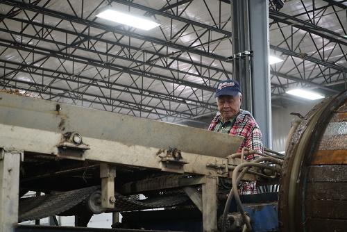 An older man wearing a baseball cap works behind a large metal machine. The machine fills the foreground with the man only visible from the chest up. 