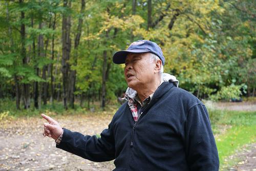 An older man wearing a baseball cap and fleece sweater stands outside facing the left of the frame with his right hand pointing in the same direction. 