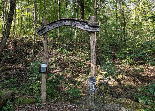 A wooden archway stands at the entrance of a trail in the woods. Carved into the top of the arch: UpS MEDICINE PLANT TRAIL.  