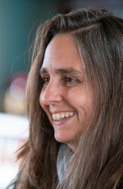 A close up of a woman’s left side profile. She has brown hair and eyes and is smiling toward the left of the frame.