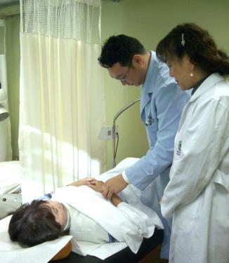 A man and woman both wearing doctor’s coats stand on the right side of the frame and look at a woman lying on an examination table. 