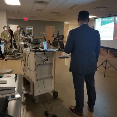 A man wearing a suit stands next to a projector cart with his back to the camera. To his right is a screen showing a slide titled “Acupuncture.” 