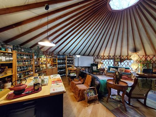The inside of this large tent features a cone-shaped fabric roof supported by wooden planks rising upward to a center skylight. The left wall is lined with wooden shelves filled with glass jars and the right side of the tent is set up like a living room. 