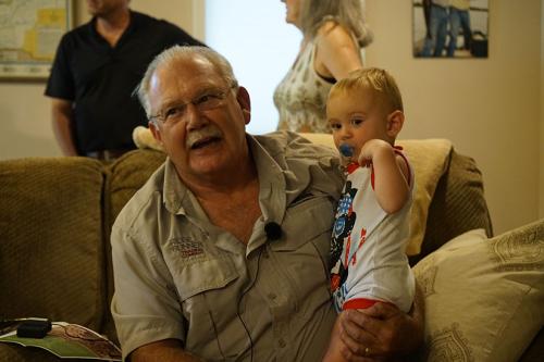 A man with gray hair sits on a couch with a baby standing on his lap. The man has a small microphone clipped to his collar. 