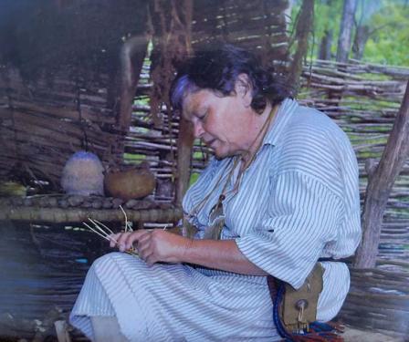 A woman sits in a wooden structure and constructs something out of a twig-like material. 