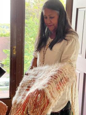 A woman with graying hair holds a woven piece of clothing featuring white feathered threads running in horizontal lines and multicolored pink threads hanging from the bottom of the piece. 