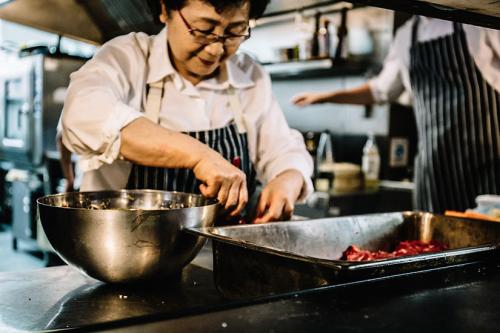 A woman wearing glasses and a striped apron in an industrial kitchen looks down at the food she is preparing. On the counter in front of her sits a large metal bowl and tray filled with unidentifiable red food. 