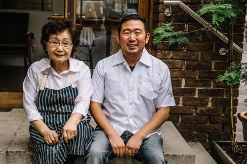 A woman wearing glasses and a striped apron sits on concrete steps in front of a building. A man wearing a white button up shirt sits to her right, our left. Both are smiling at the camera. 