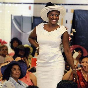 Black woman poses in form-fitting white dress and white church hat, as other seated women in hats look up and take photos.