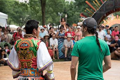 Two people facing away, one in a T-shirt and the other in a brightly embroidered vest, toward a crowd of people outdoors.