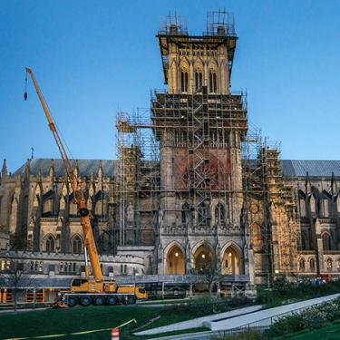 Against a dusky dark blue sky, the National Cathedral stands blocked by extensive scaffolding and a tall yellow crane.