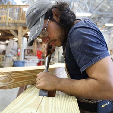 A young man fine tunes a cut in a piece of lumber using a hand tool that looks like a wide chisel.