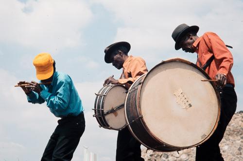 Three men form a line while playing music. The one in front plays a fife, and the two behind him play drums strapped to their chests. Old color photo.