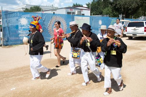 Four men in matching black shirts and white pants, and a young woman in bright red shirt and shorts, march down a gravel pathway, with a sign for the Smithsonian Folklife Festival behind them. Each person holds or plays an instrument: a conch, flutes, vio