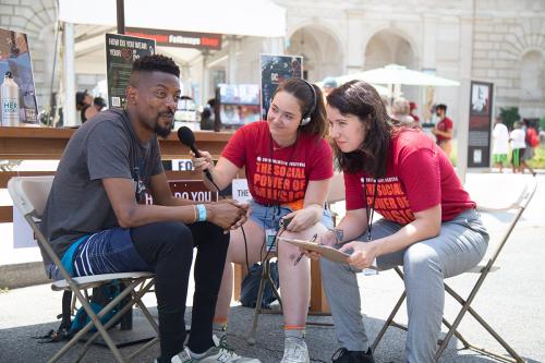 Seated in folding plastic chairs, two young women in matching red staff shirts interview a man across from them, one holding a microphone to his mouth as he smirks and looks in the distance. Outdoor festival tents and visitors in the background.