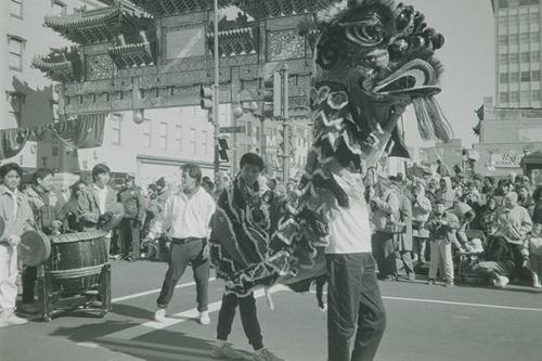 Two young men enliven a Chinese dragon costume, the the one in front raising its head high. Behind them, other young men play percussion instruments, and a crowd forms under the Chinese archway over the street.