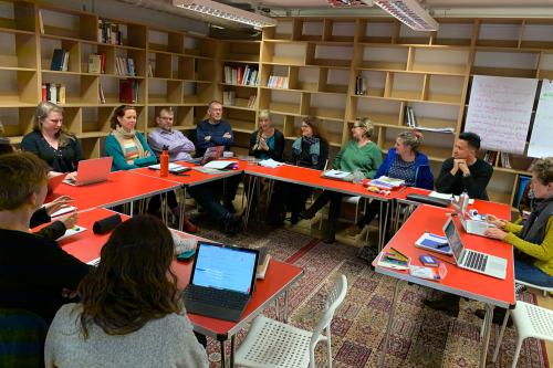 A group of people with laptops in a conference room filled with bookcases having a discussion.