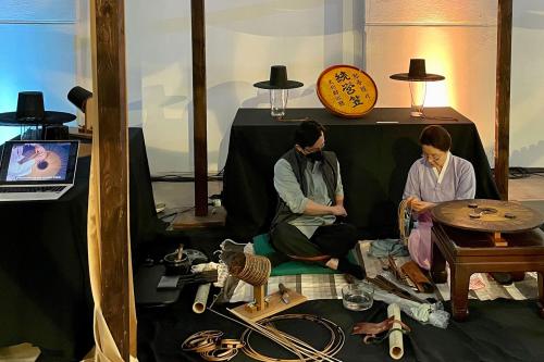 An older woman and a young man sit next to eachother on a floor. The woman, in front holds a flat wooden disk in the shape of a hat to bend horsehair around.