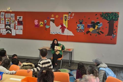 An adult sits in a classroom while reading a book. Children sit on a rug and listen.